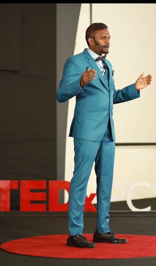 Fatha Khan speaking on the TEDx stage in 2022 in a teal suit